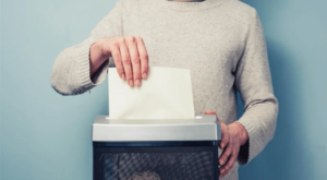 An image of a person shoulders down in front of a plain blue background. They have a shredder in front of them holding up a piece of paper in their hand as it is shredded by the machine.